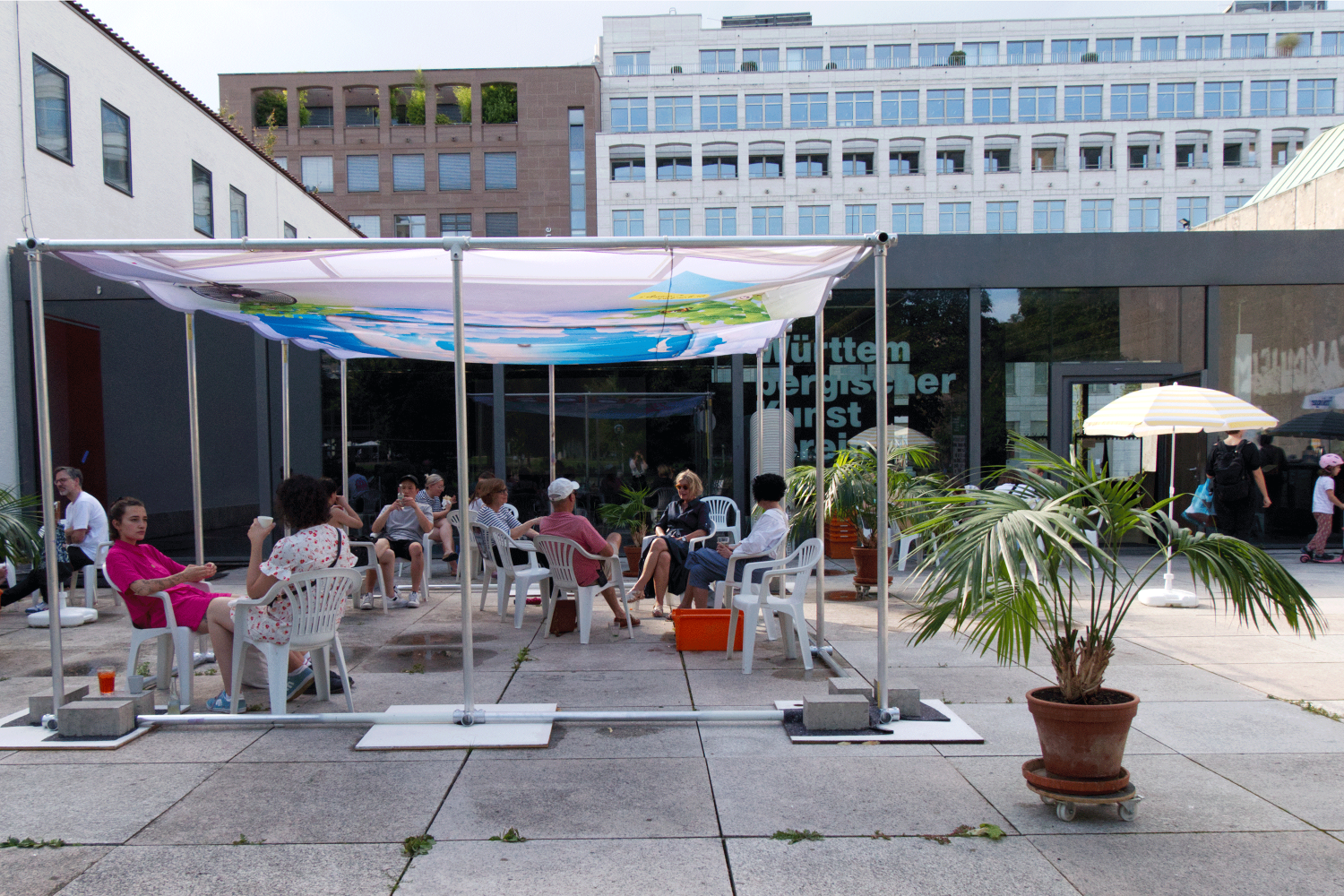 Menschen sitzen auf weißen Monoblocs unter dem Sonnensegel “Sky Notes” der Künstlerin Victoire Gonzalvez.