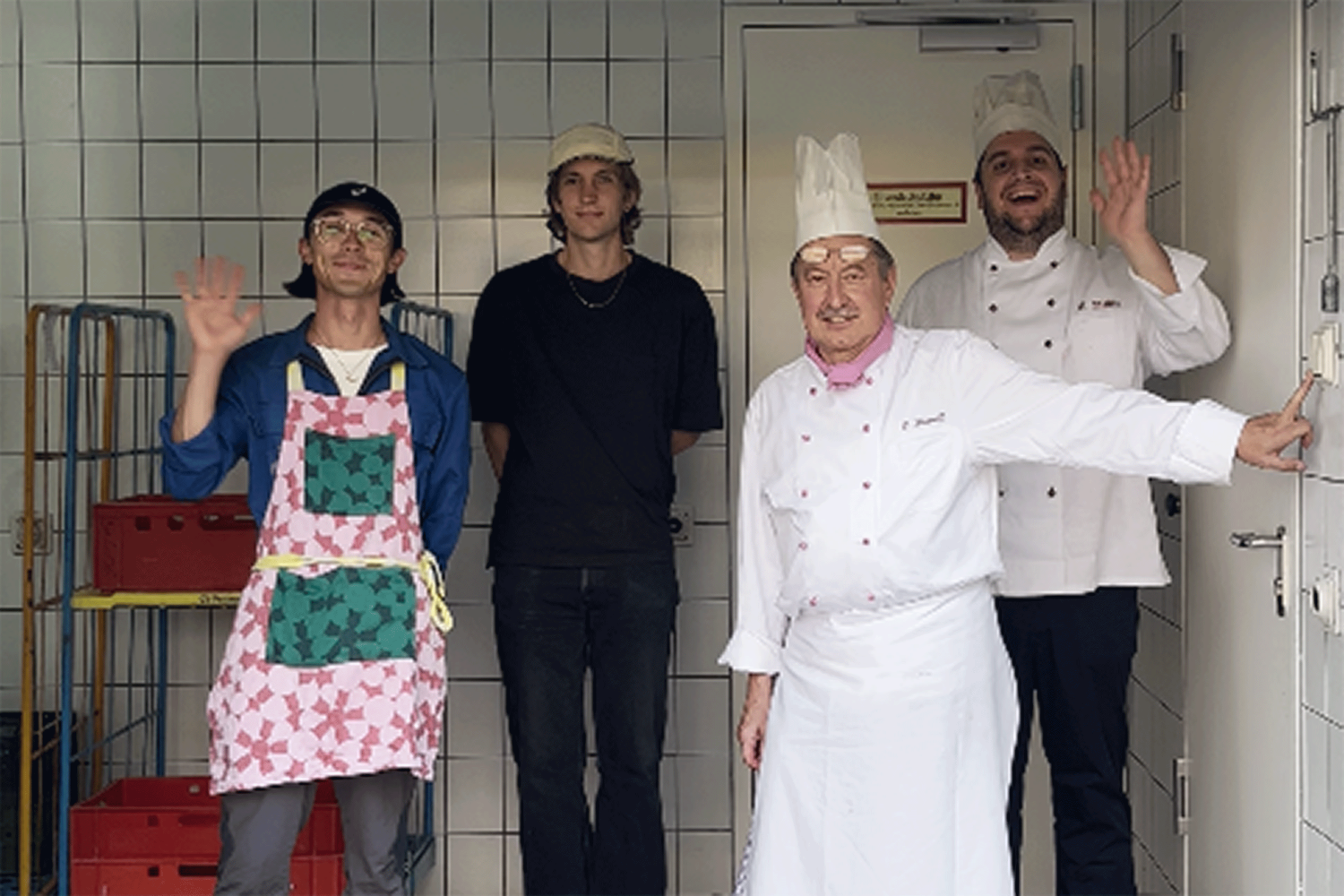 Hinter einem aufgehenden Rolltor stehen vier winkende Personen. Zwei davon tragen klassische Kochkleidung und -mützen. Die anderen beiden tragen legere, bunte Alltagskleidung mit Schürze.
