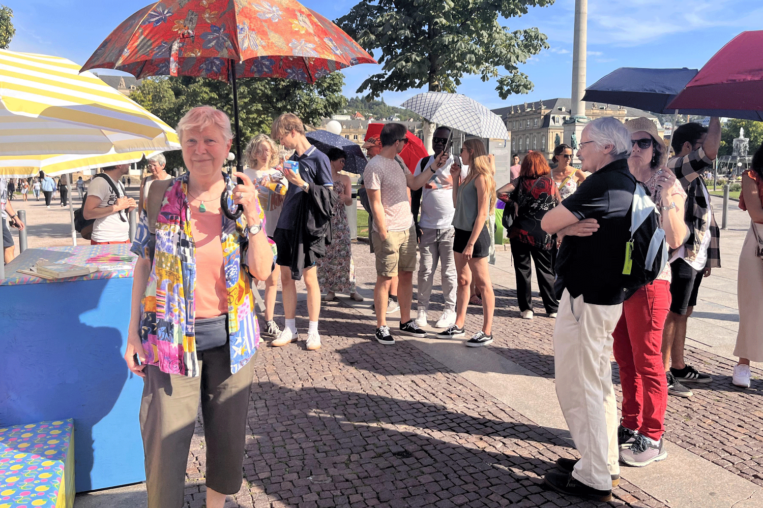 Im linken Bilddrittel steht die Stadtökologin Barbara Drescher mit knallbuntem Sommerhemd und Schirm. Hinter ihr versammeln sich die Teilnehmer*innen und warten auf den Beginn des Stadtspaziergangs.